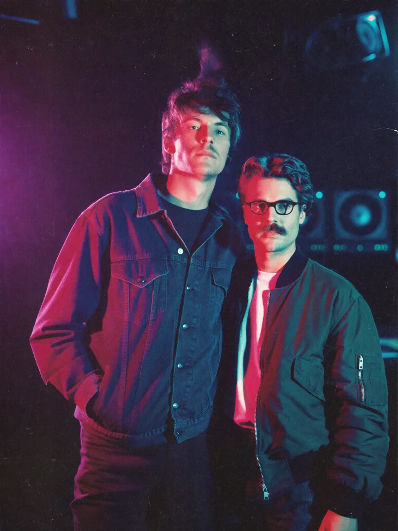 Promo photograph of Vector Hearts: a duo lit by magenta and cyan light in a dark television studio